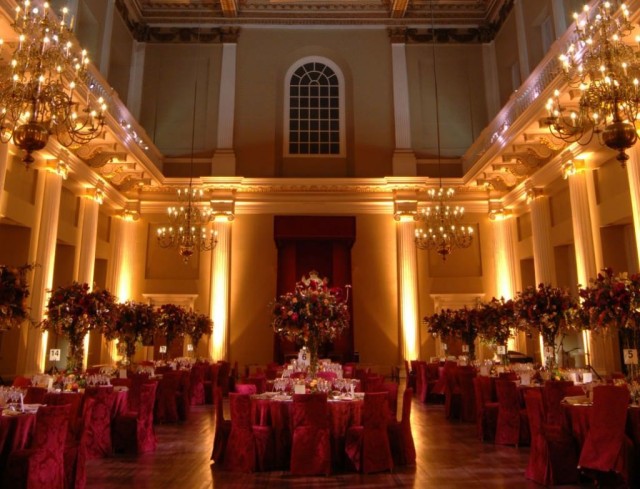 Main Hall set for a formal dinner with round tables decorated with large floral centre pieces, the high ceilings are accentuated by the uplighters Banqueting House Venue Hire SW1